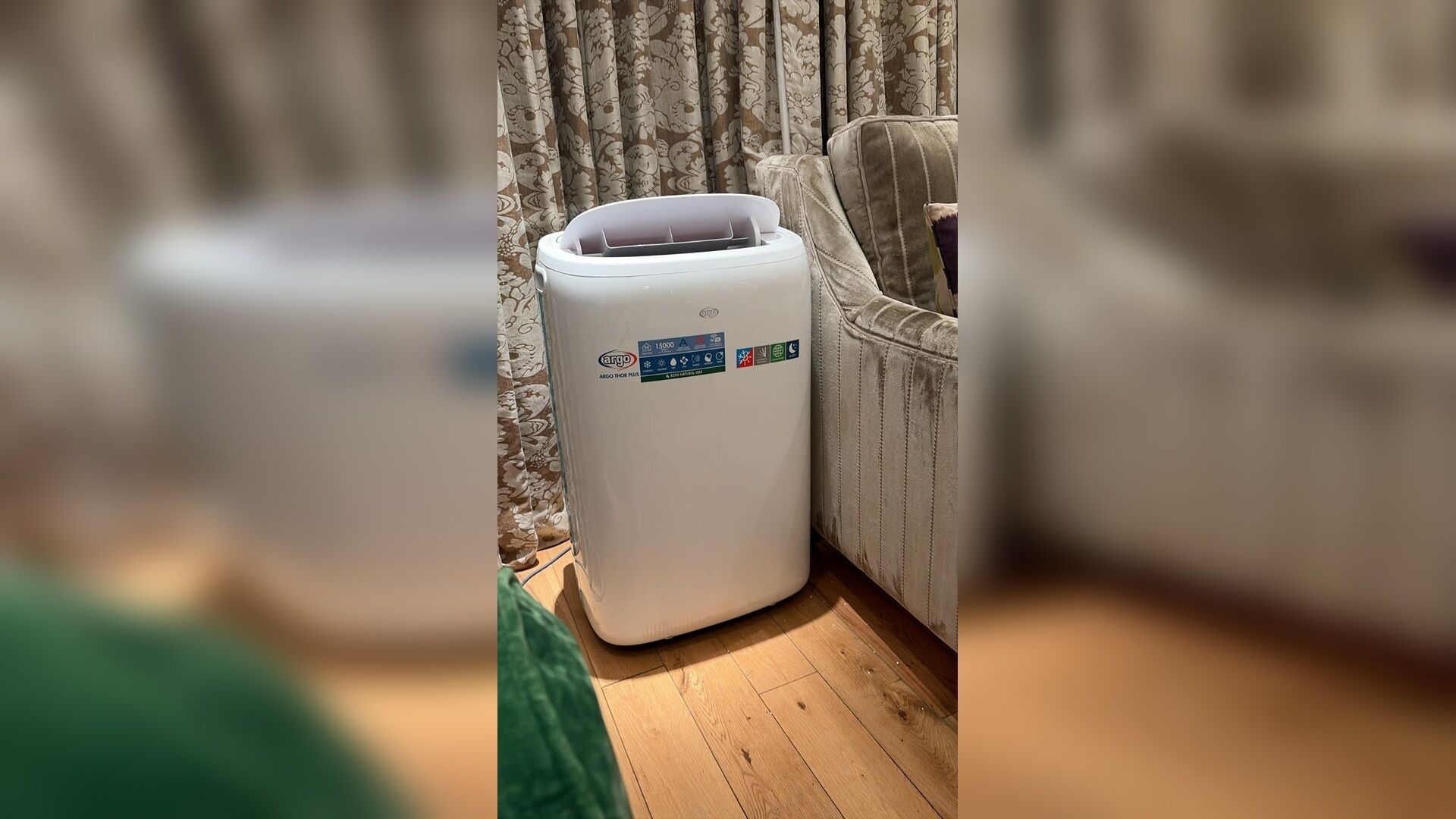 A white portable air purifier with a digital display and certification labels, placed on a wooden floor next to a sofa with a patterned curtain in the background.