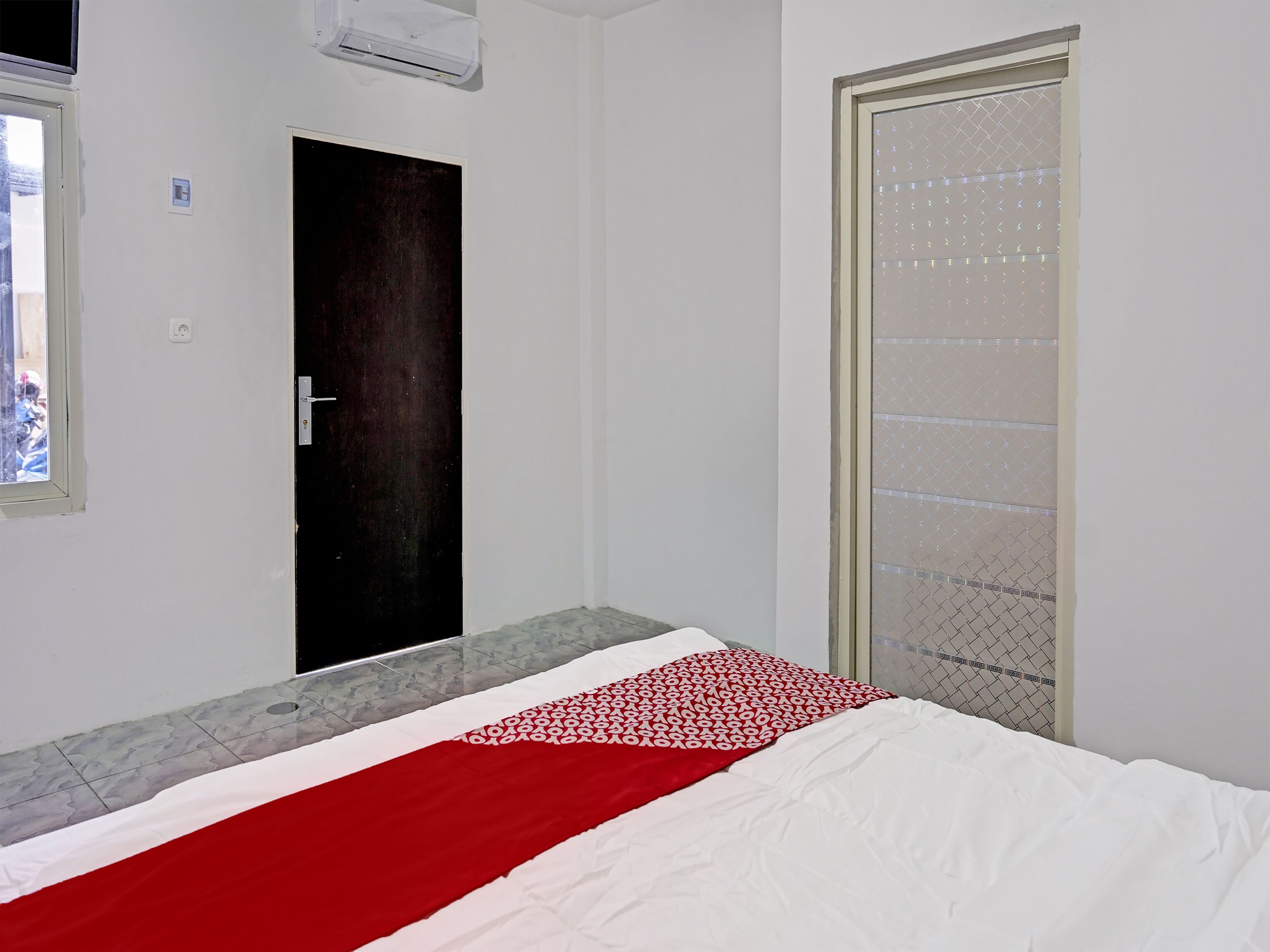 A room with a bed, a dark door, a white wall, and a window.