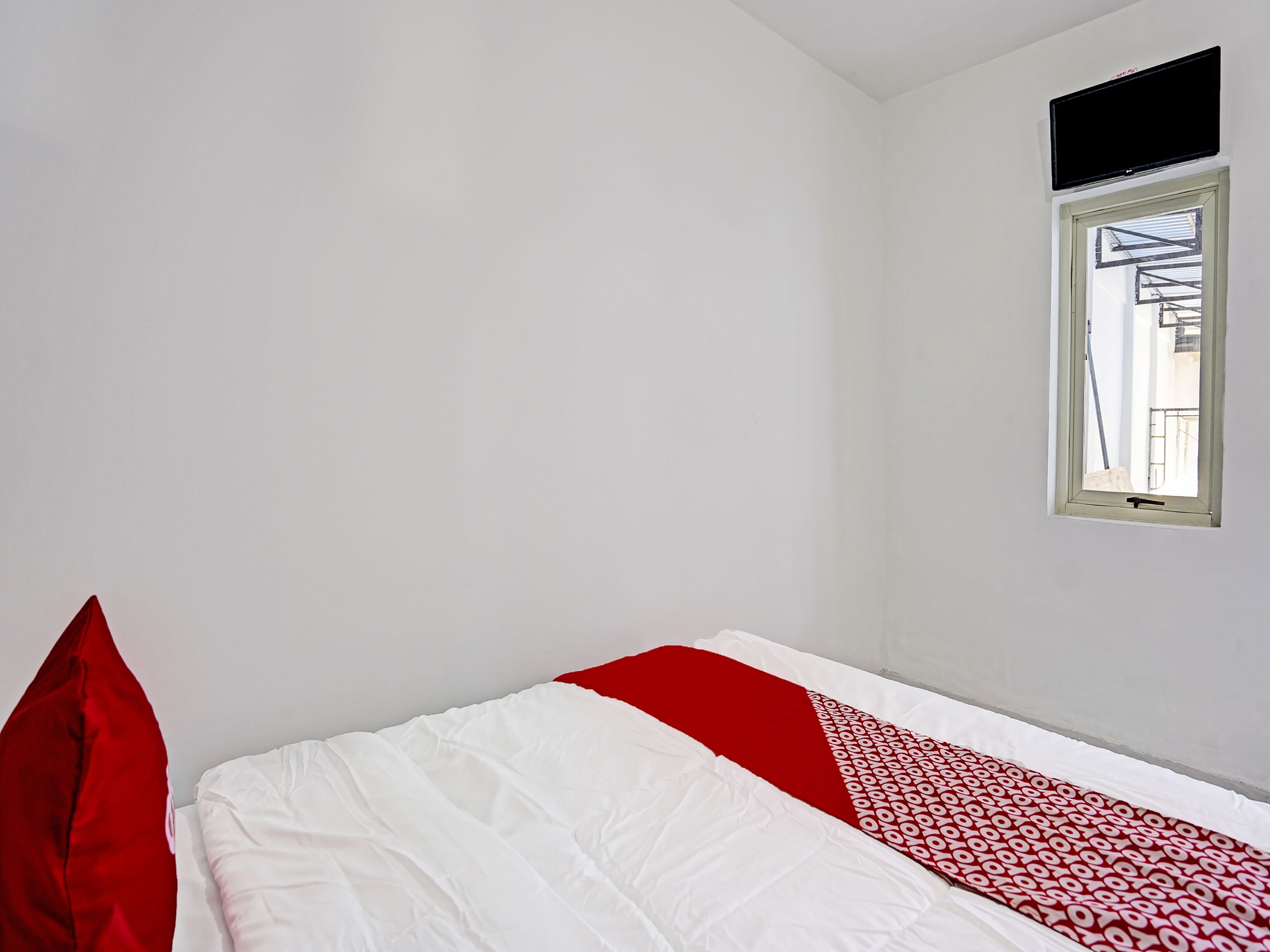 A small, sparsely furnished room with a single bed, a window, and a television mounted on the wall.