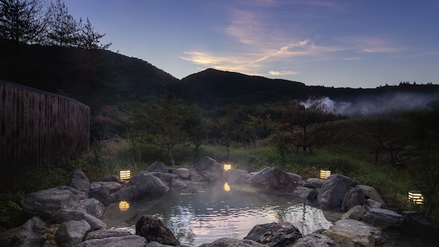 A hot spring surrounded by rocks and vegetation, with a wooden structure on the left and lanterns along the edge. - Hoshino Resorts KAI Unzen (Unzen)