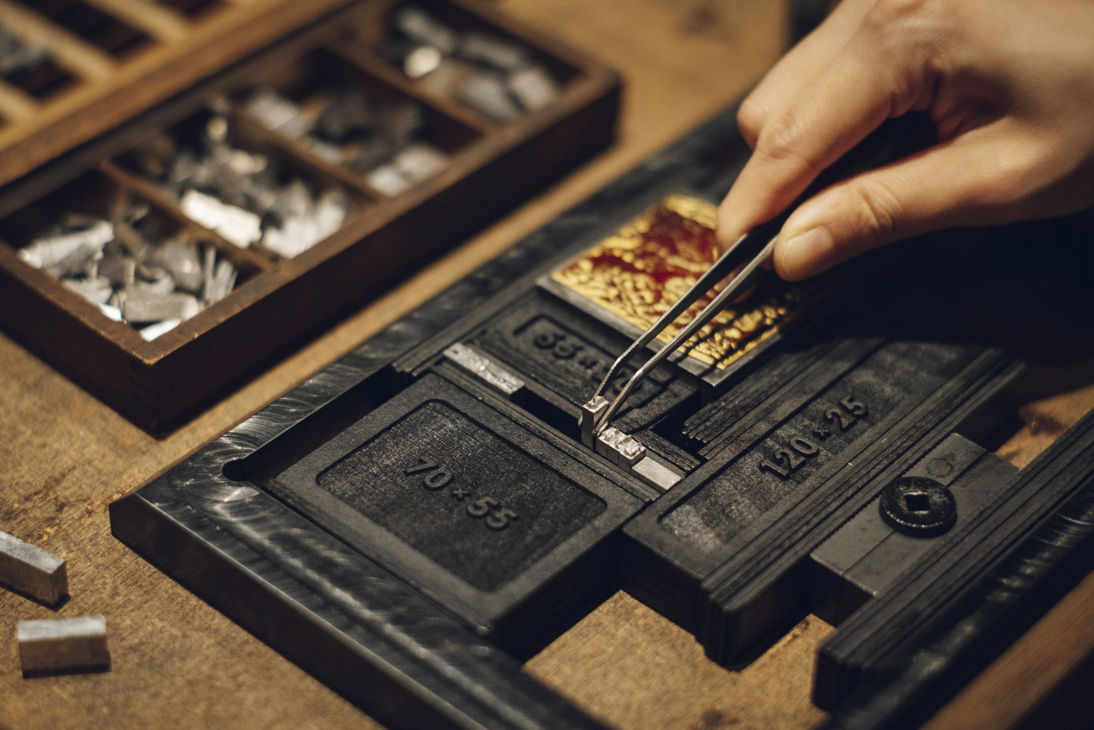 A close-up of a hand using a tool on a metal object, with a wooden box containing various metal parts in the background.