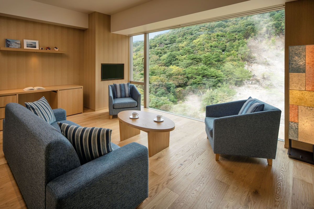 A modern living room with a large window, a sofa set, a coffee table, and a flat-screen TV.