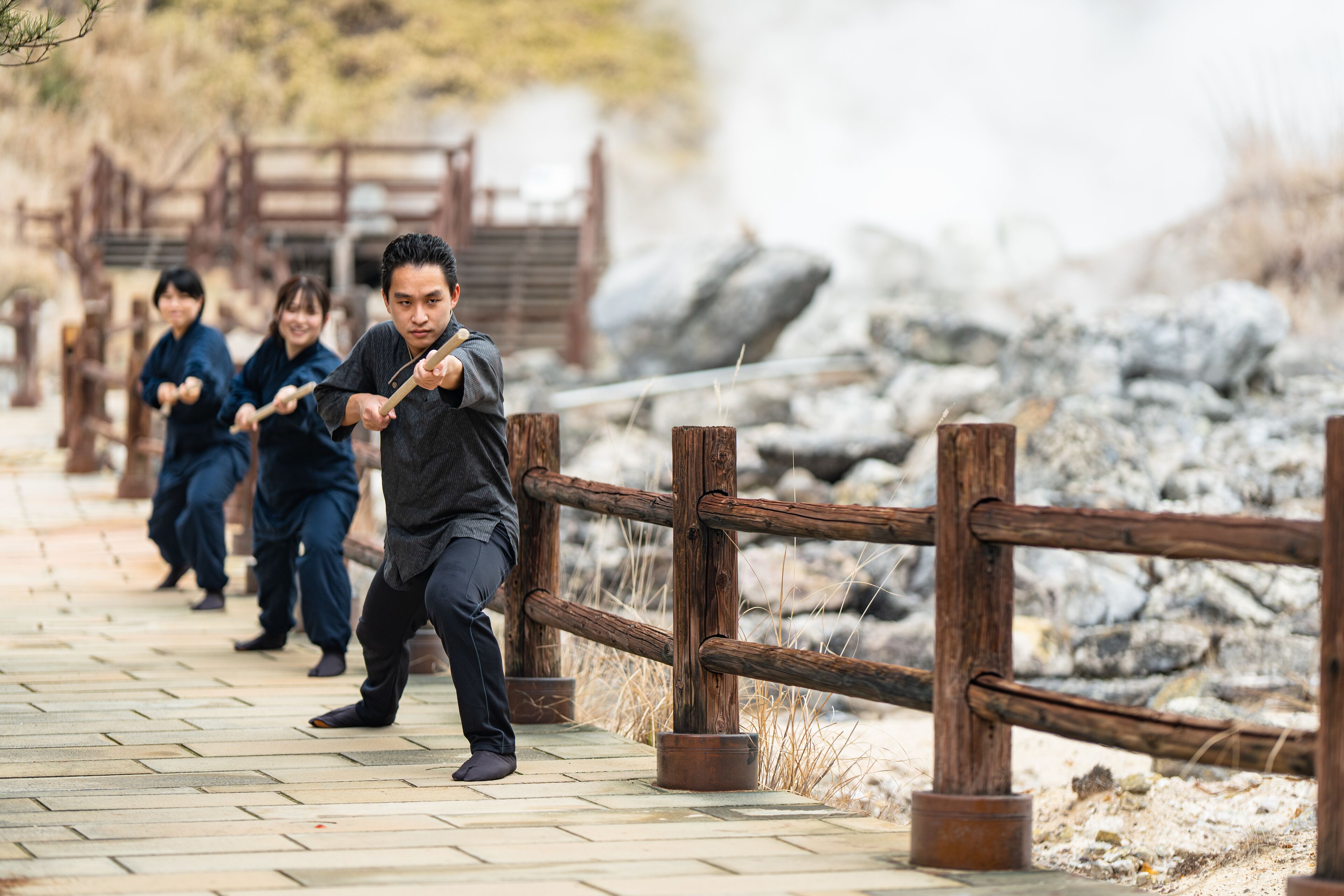 A group of people practicing martial arts on a paved path with wooden railings and a rocky background.