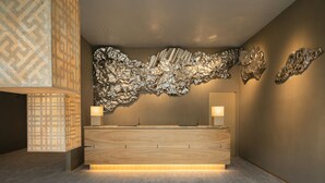 A modern reception area with a wooden reception desk, two wall-mounted lamps, and a large abstract metallic wall art piece. - Hoshino Resorts KAI Unzen (Unzen)