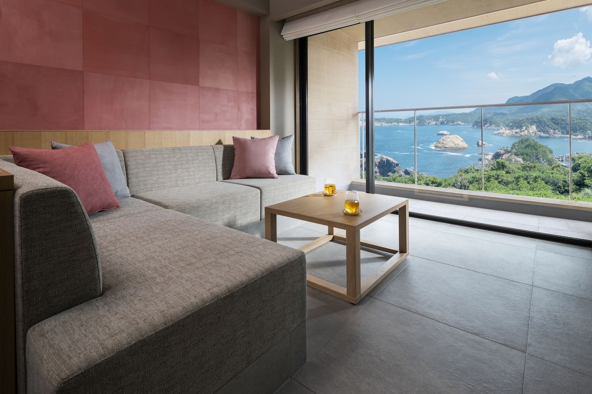Sala de estar moderna com um sofá cinzento, uma mesa de centro de madeira e uma grande janela com vista para o mar e montanhas.