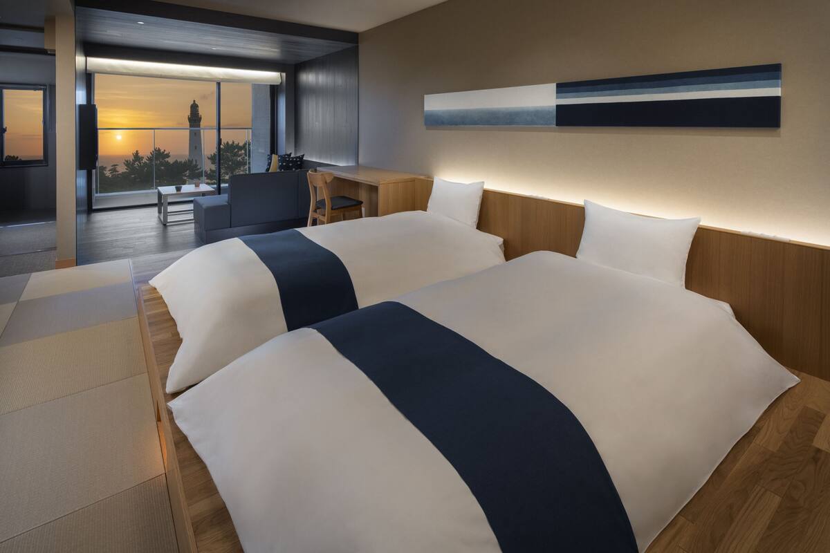 Quarto de hotel moderno com duas camas, uma escrivaninha e vista para o pôr do sol através de grandes janelas.