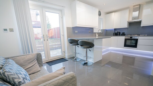 A modern kitchen with white cabinets, a built-in oven, and a coffee maker. There is a seating area with a sofa and two bar stools. - Newly Refurbished Modern 4 Bed Detached House (oxford)