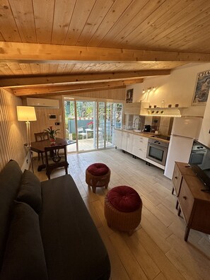 A cozy wooden interior with a sofa, two red poufs, a dining table with chairs, and a kitchen area with a fridge, oven, and sink. - The Little House (Roma)