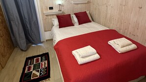 Une chambre avec un plafond en bois, un lit avec une literie rouge et blanche, une petite table de chevet avec une lampe, et un tapis à motifs sur le sol.