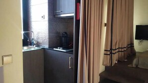 Een compacte keuken met een inbouwfornuis, een koffiezetapparaat en een spoelbak. Er is een zithoek met een bank en een aan de muur bevestigde televisie.