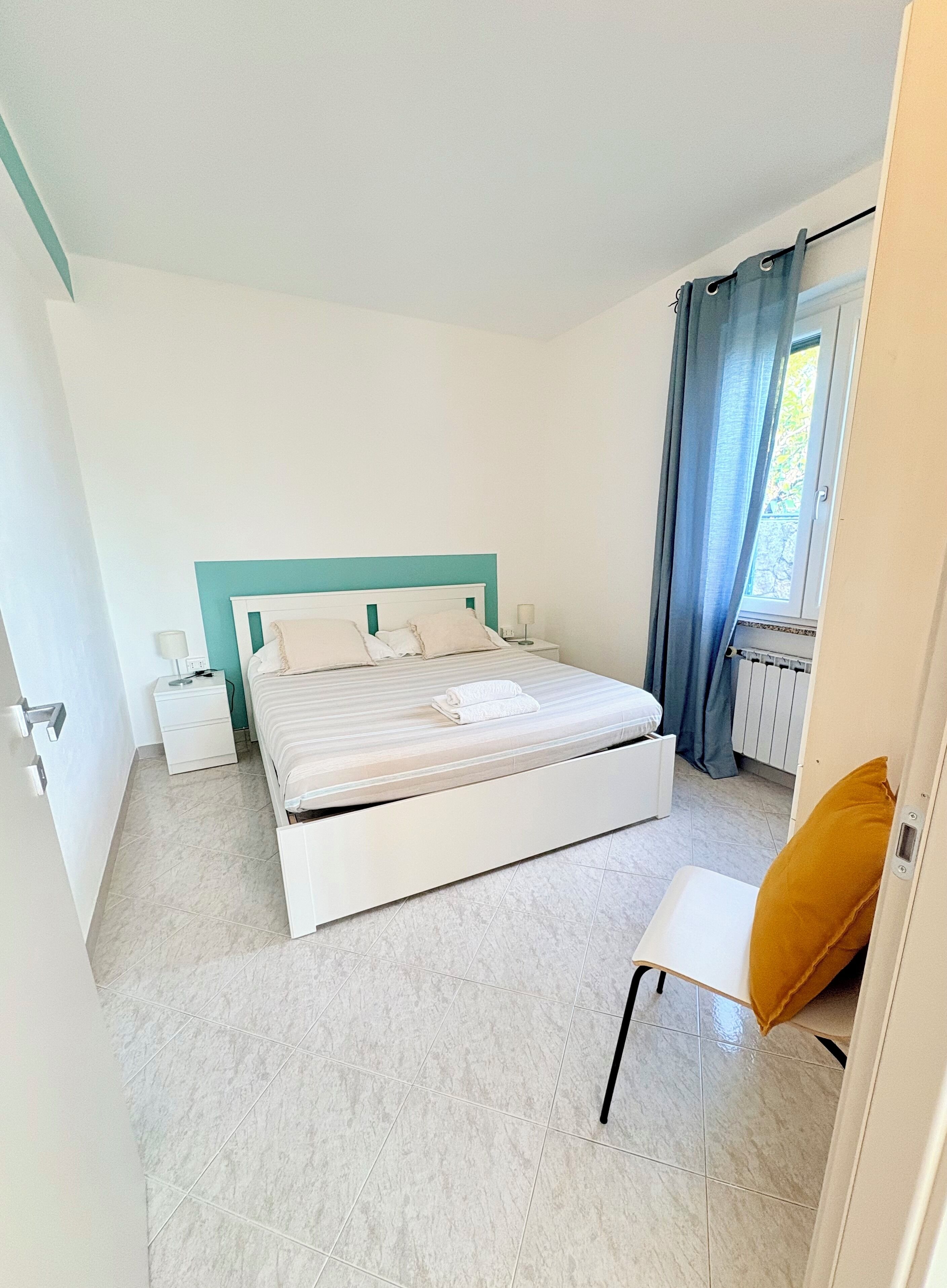 Un dormitorio con cama, mesita de noche, silla y ventana con cortinas.