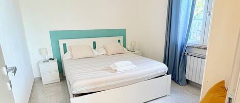Un dormitorio con cama, mesita de noche, silla y ventana con cortinas.