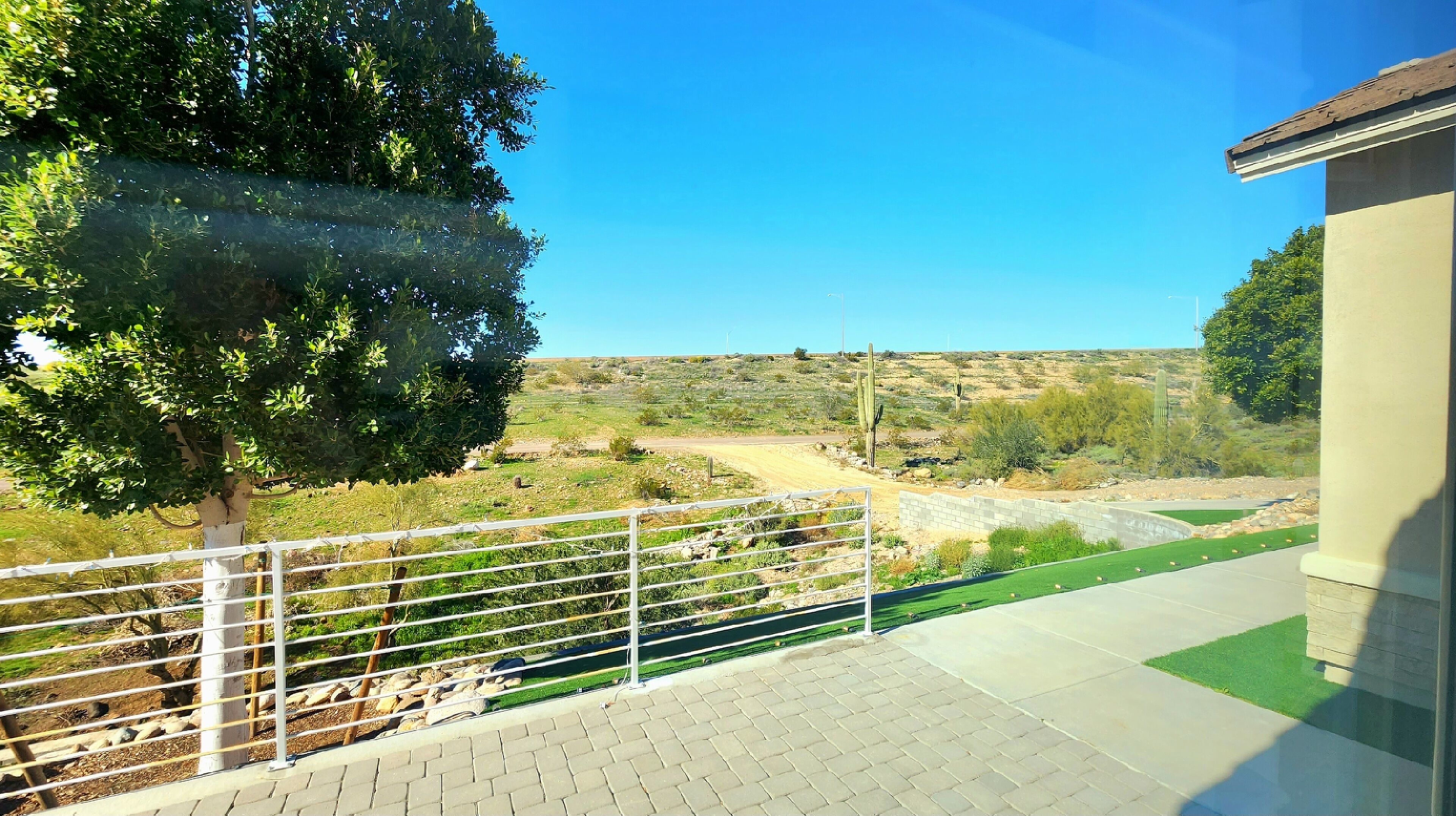 A paved balcony with a metal railing overlooking a desert landscape with sparse vegetation and a dirt road.