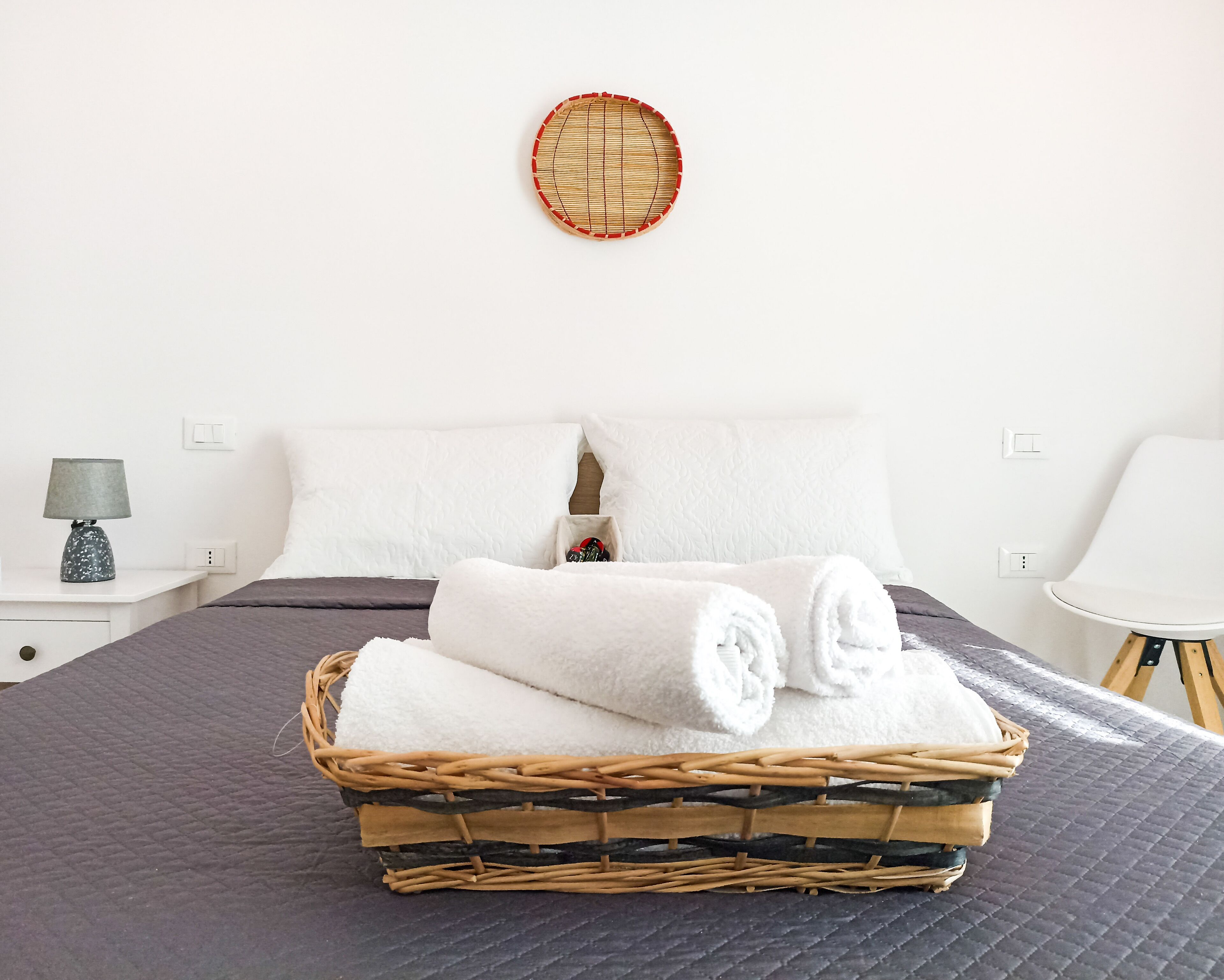 A neatly made bed with white linens, a wicker basket with folded towels, and a round woven wall decoration.