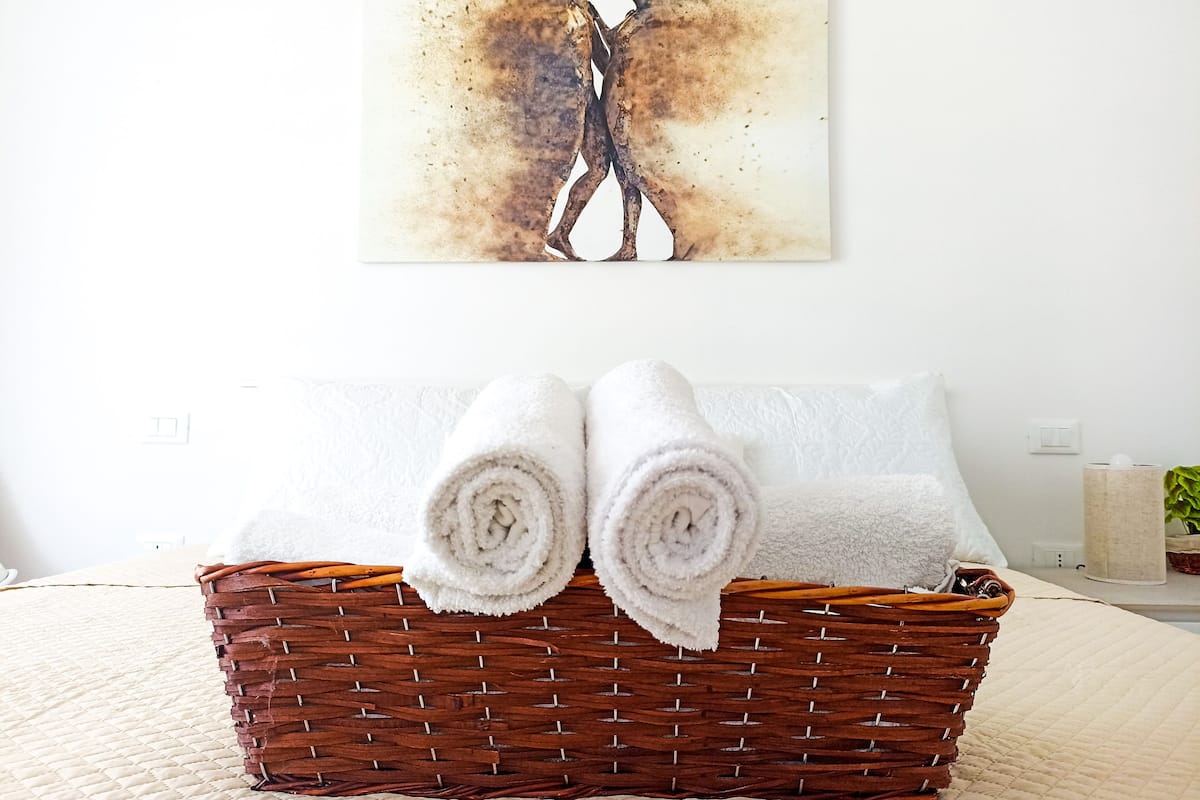 A neatly made bed with white linens, a wicker basket with rolled towels, and a painting of two figures on the wall.