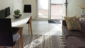 A modern living room with a dining area, a TV mounted on the wall, a ceiling light fixture, and a sliding glass door leading outside. - Kolossoi Guest House (Cabras)