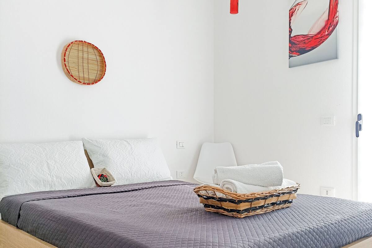 A neatly made bed with a dark quilt, a wicker basket on the bed, and a wall clock.