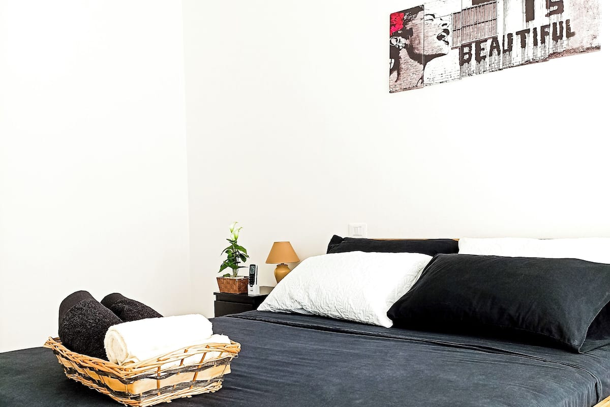A neatly made bed with a dark bedspread, a basket of towels, and a wall art piece.