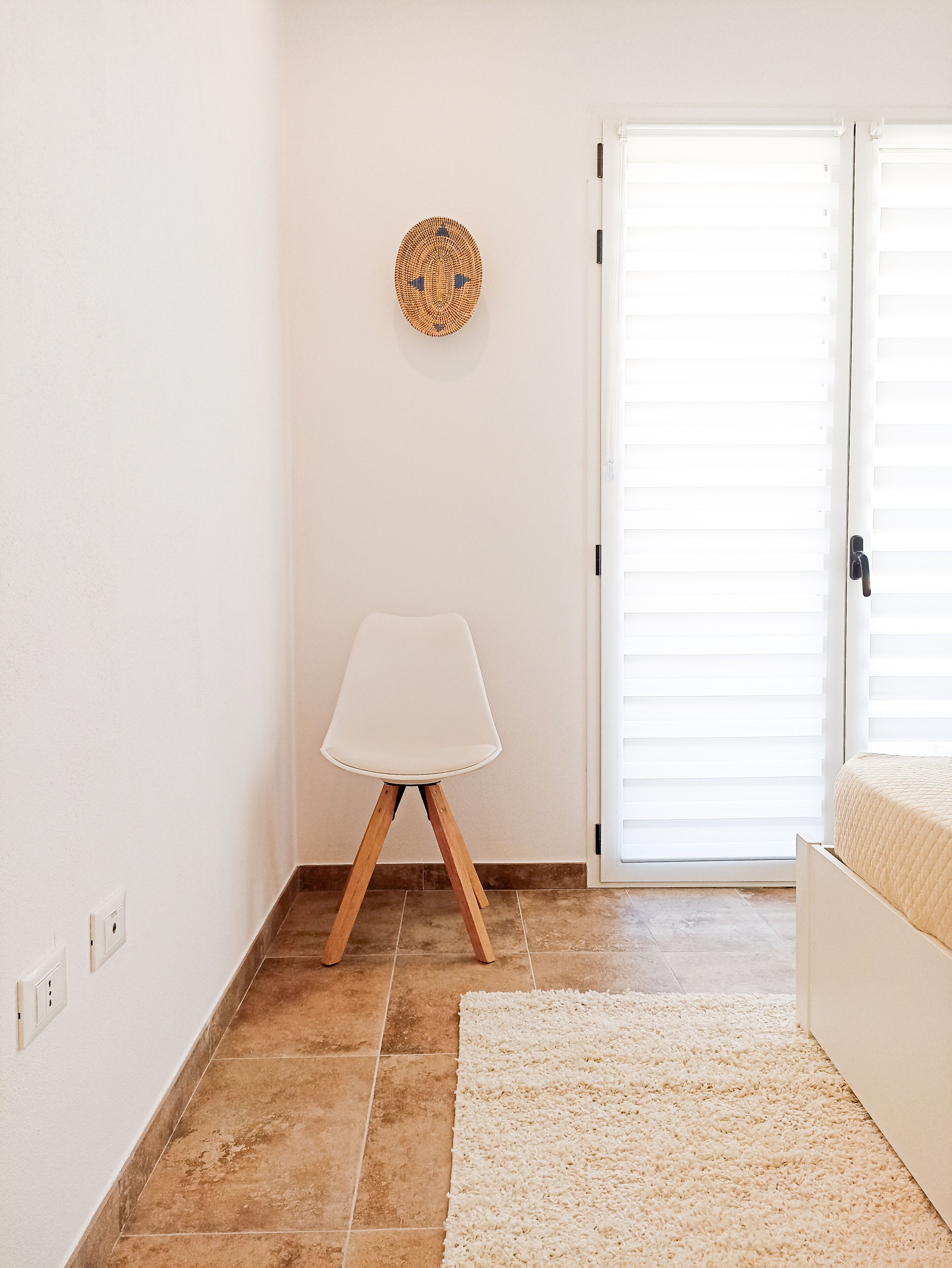 Ein Zimmer mit einer weißen Tür, einem weißen Stuhl mit Holzbeinen, einem gewebten Wandbehang und einem beigefarbenen Teppich.