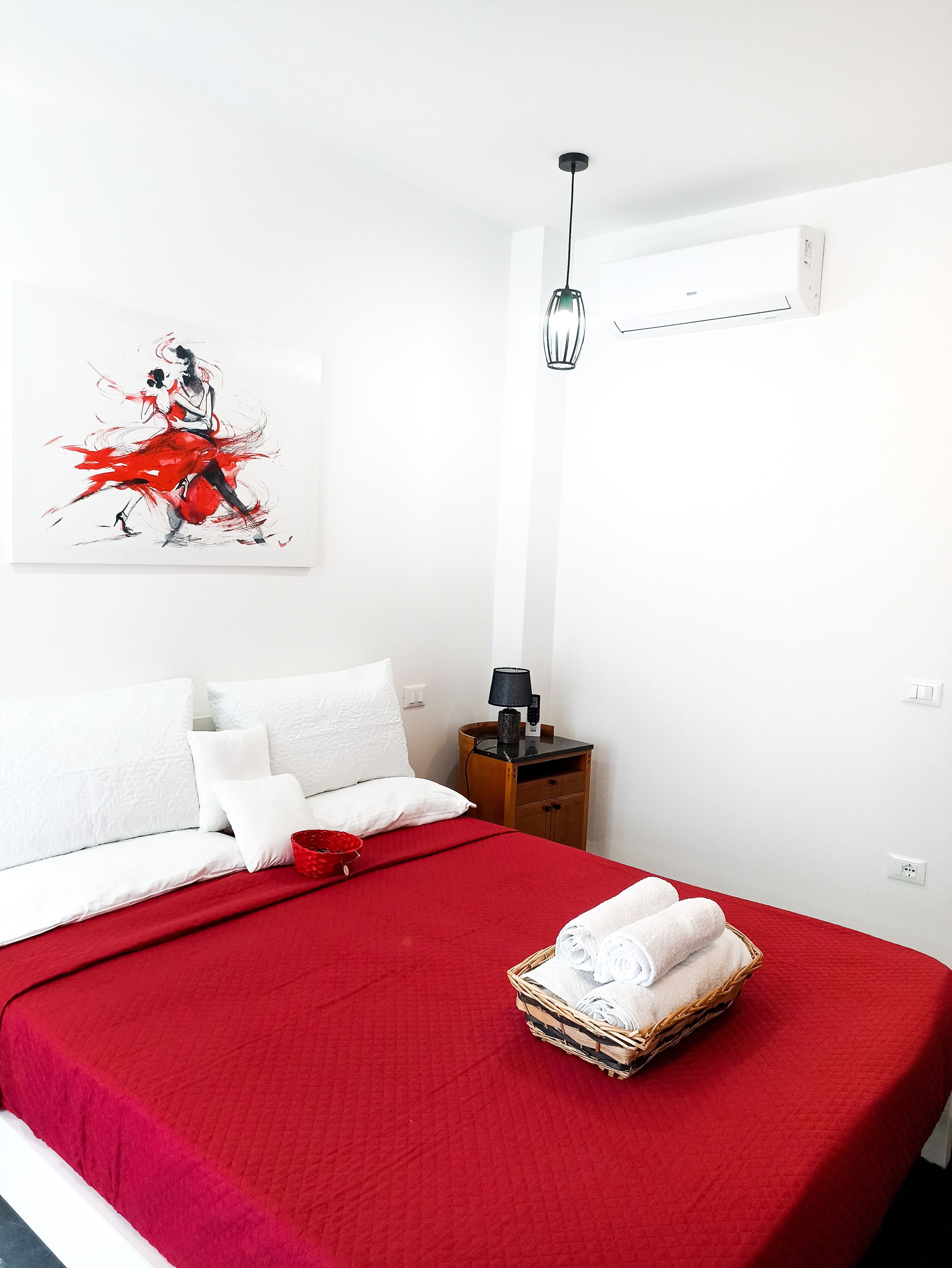 A hotel room with a bed, a red bedspread, white pillows, a white wall, a painting, a nightstand, a lamp, and a basket of towels.
