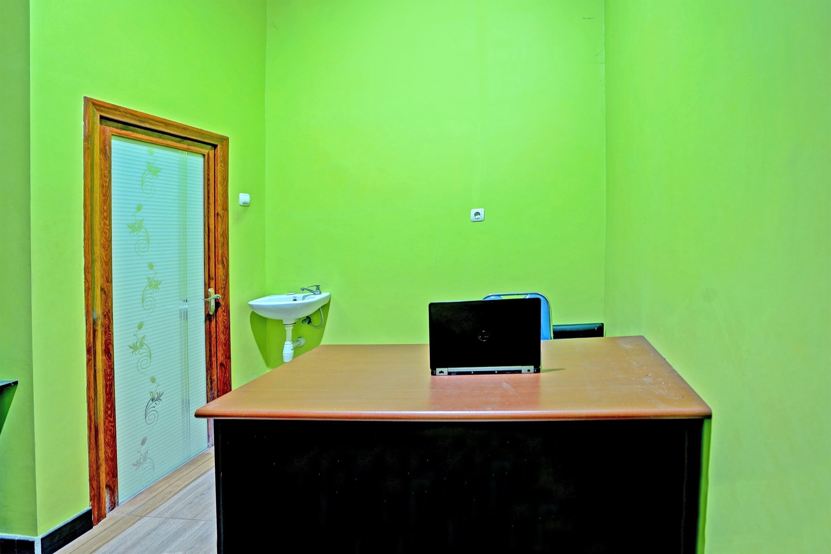 A room with a green wall, a wooden door with a glass panel, a white sink, and a laptop on a desk.