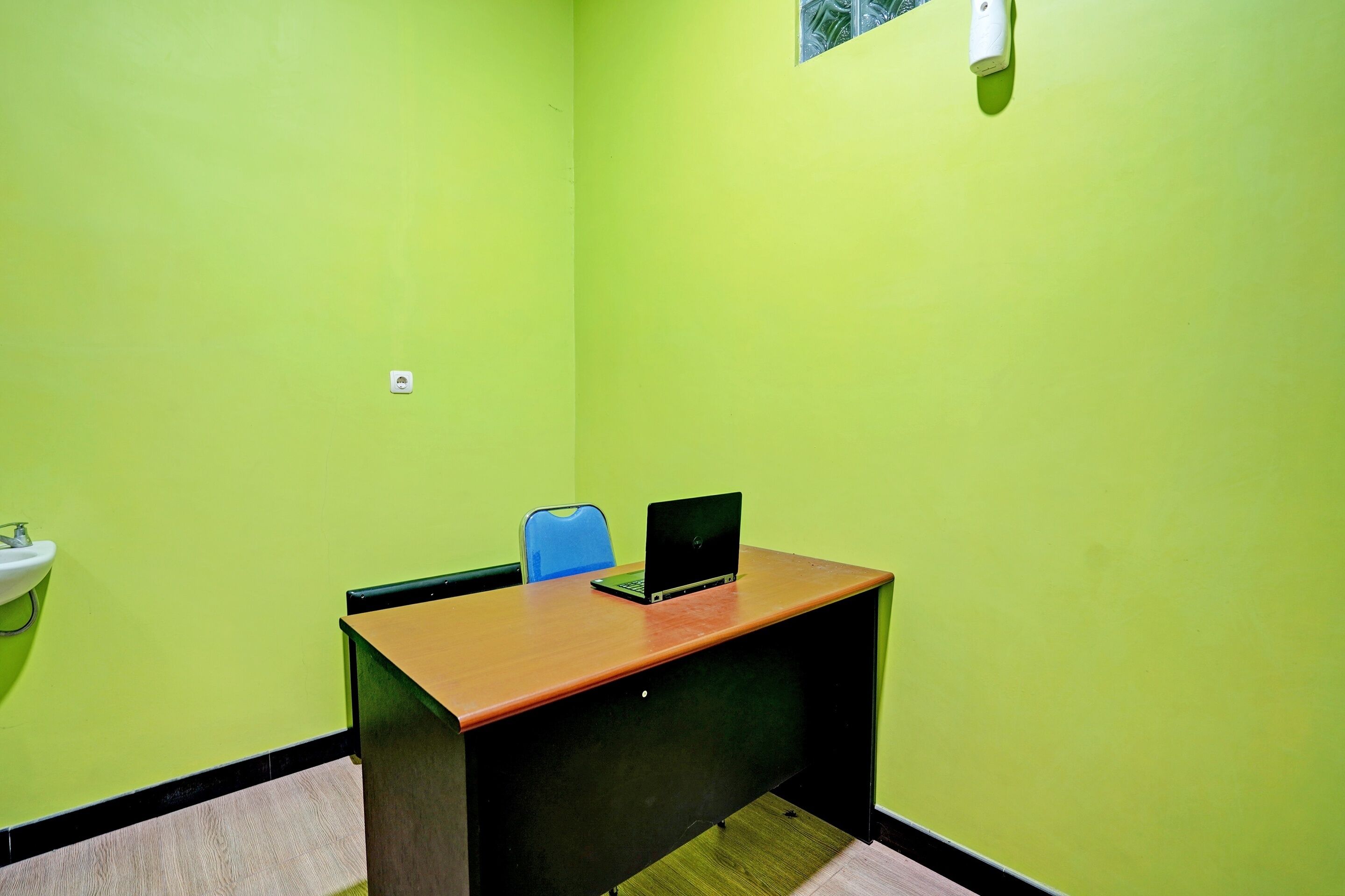 Una habitación con pared verde, un escritorio de madera con una computadora portátil, una silla azul y un lavamanos blanco.