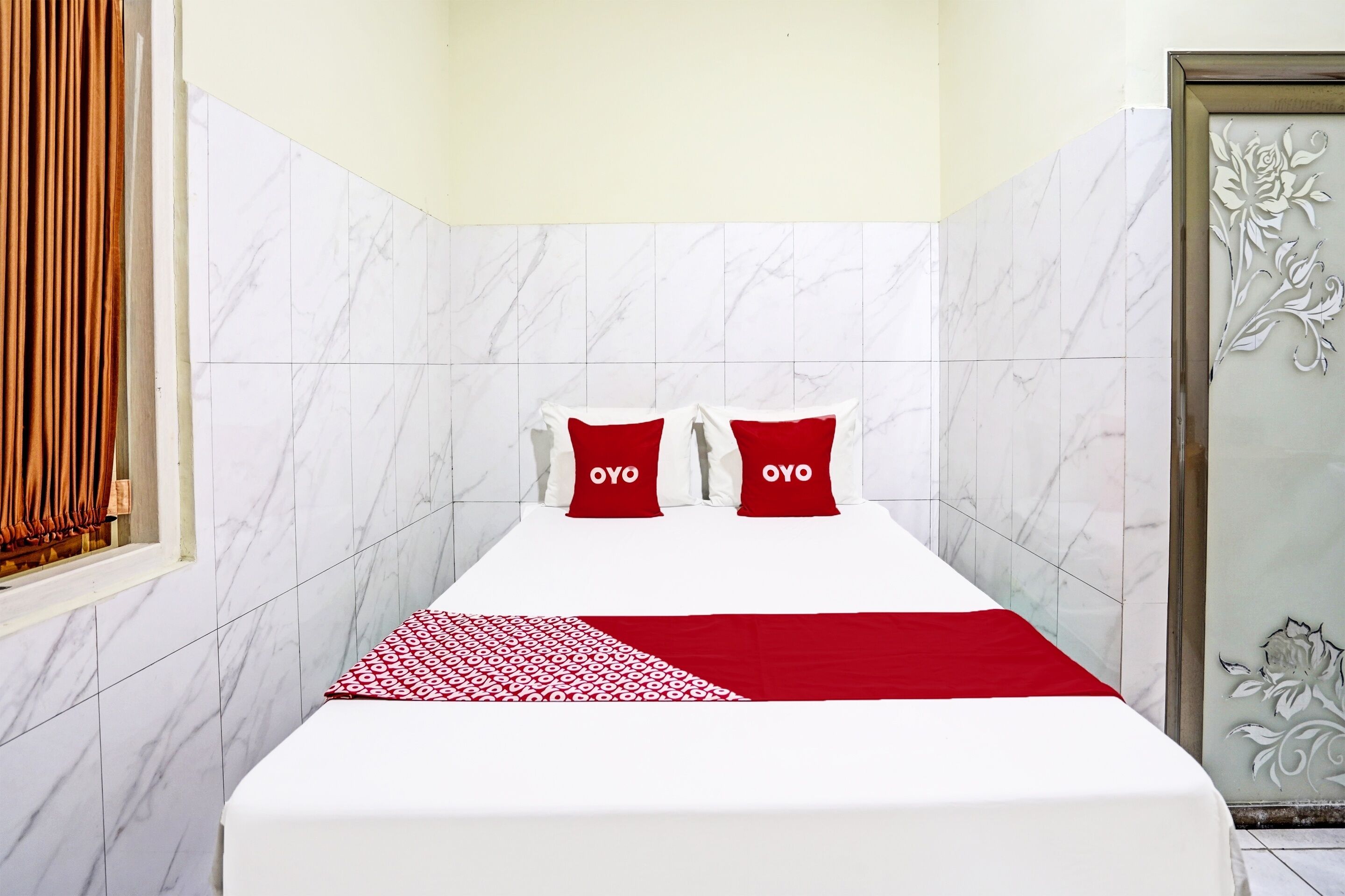 Hotelkamer met een bed, twee rode kussens met de tekst 'OYO', een rood-wit gestreept plaid en marmeren muren.