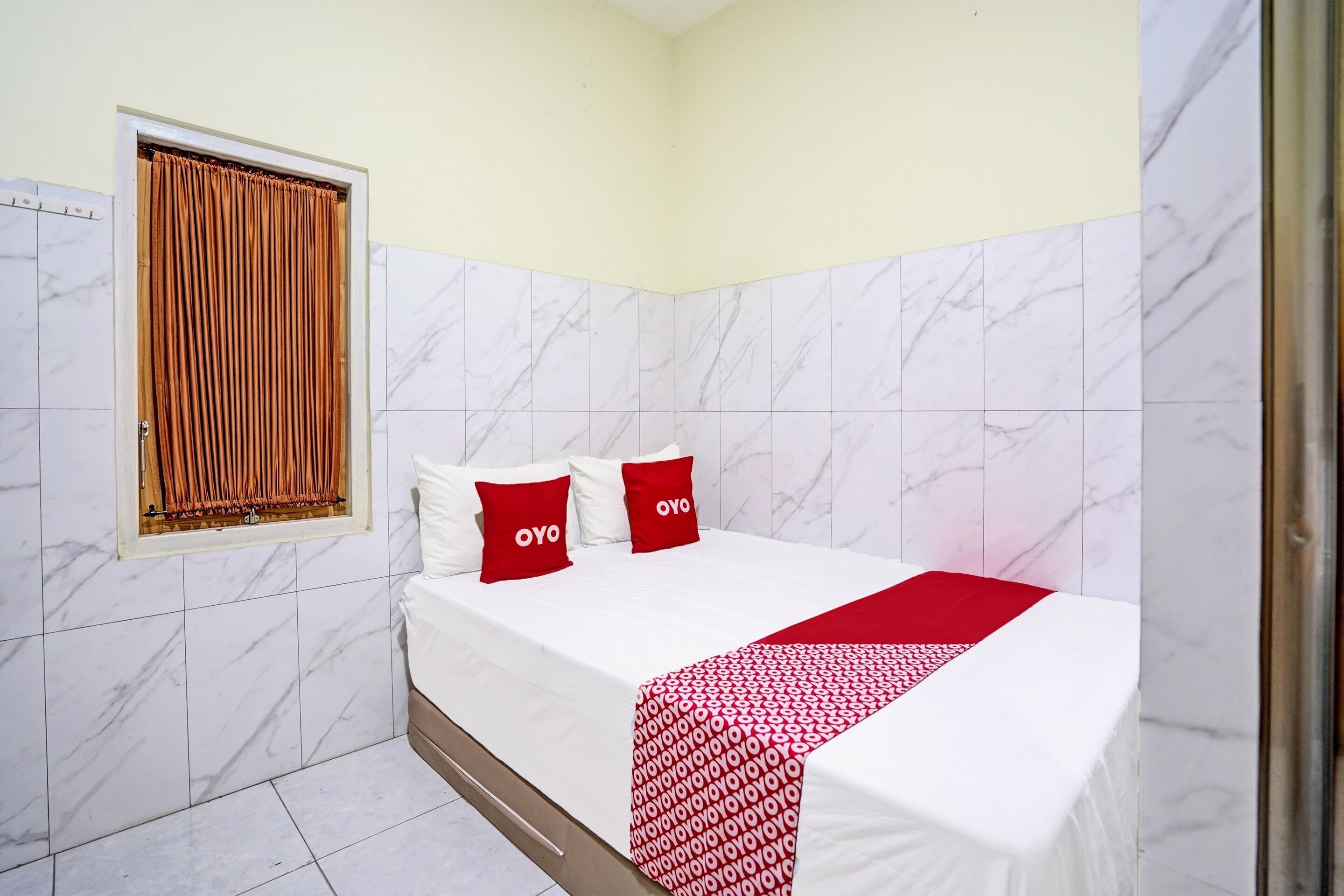 A hotel room with a bed, two pillows with "OYO" branding, a window with curtains, and marble-tiled walls.