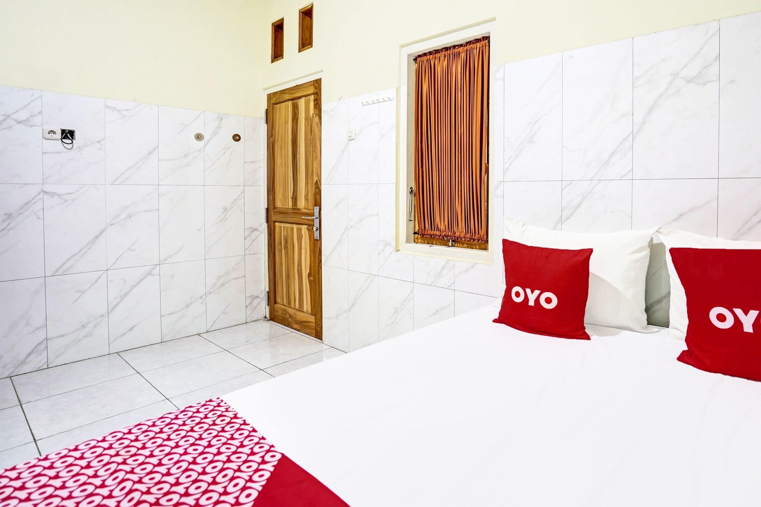 A hotel room with a bed, two pillows with "OYO" branding, a wooden door, a window with curtains, and marble-patterned walls.