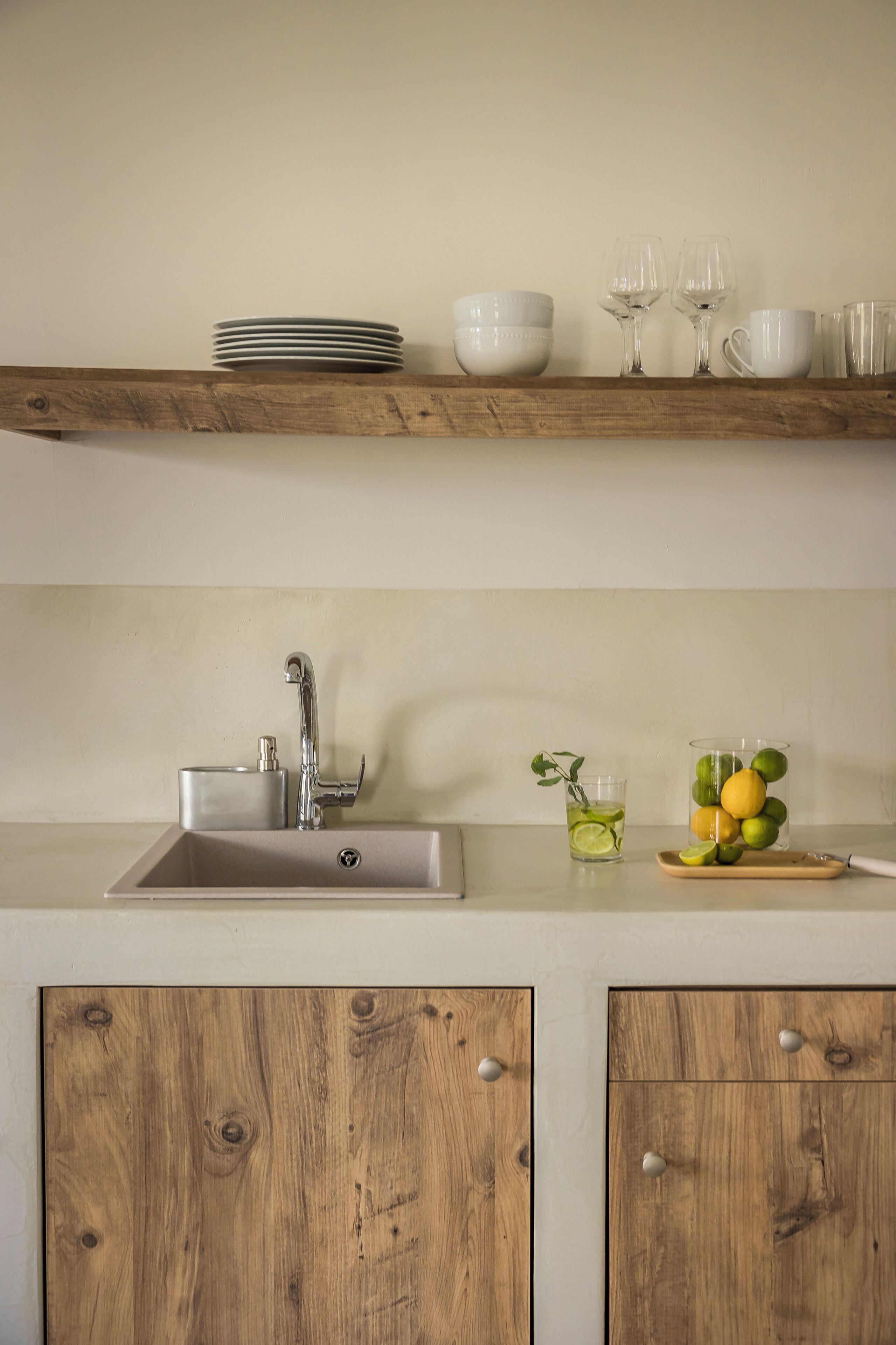 A kitchen with a wooden shelf holding plates and glasses, a sink with a faucet, and a fruit bowl with lemons and limes.