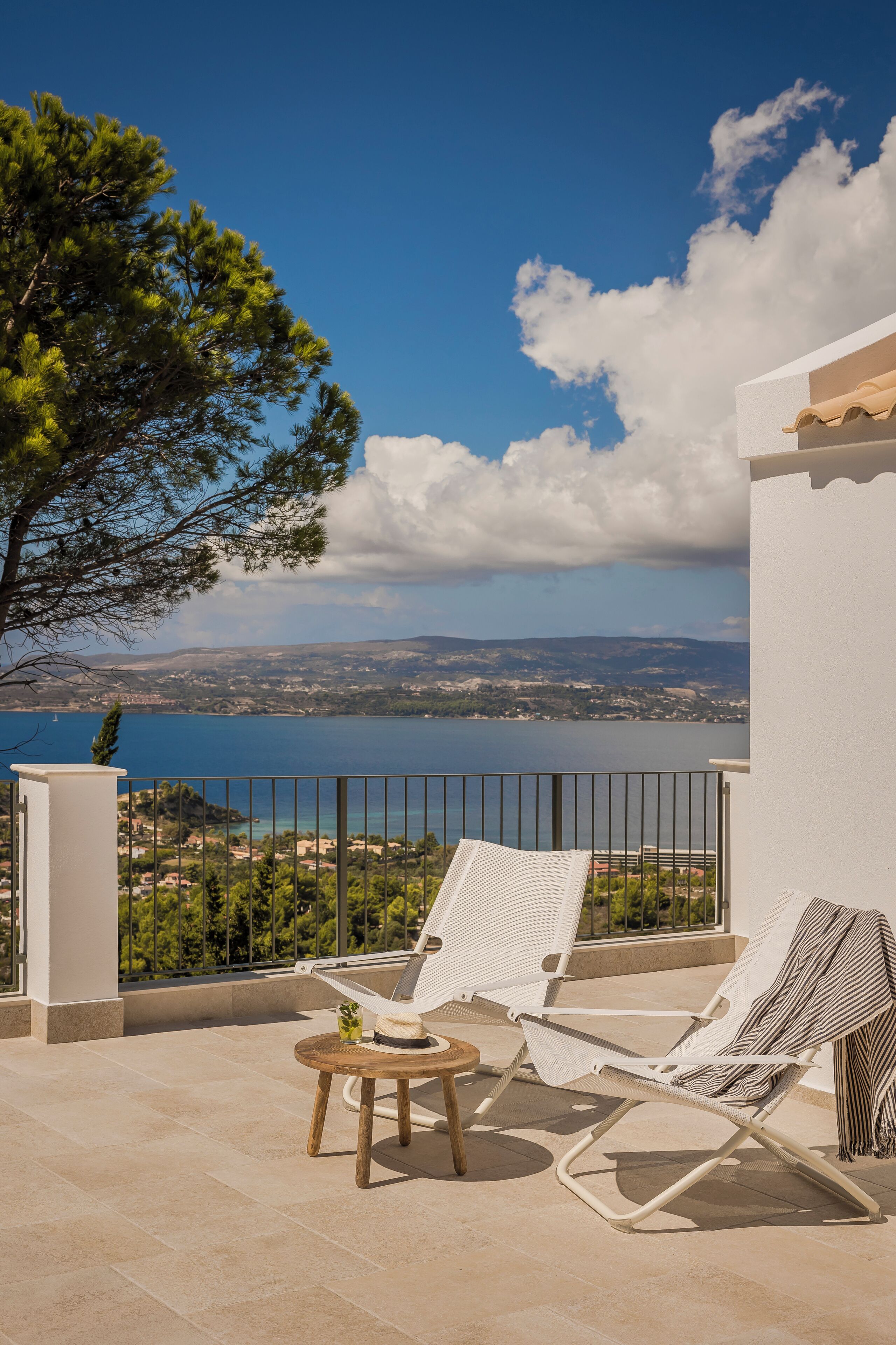 A balcony with two white lounge chairs, a small round table, and a view of a lake and distant hills.