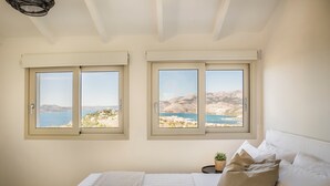 Un dormitorio con una cama, dos ventanas que dan a un paisaje costero y una planta pequeña en una mesita de noche.