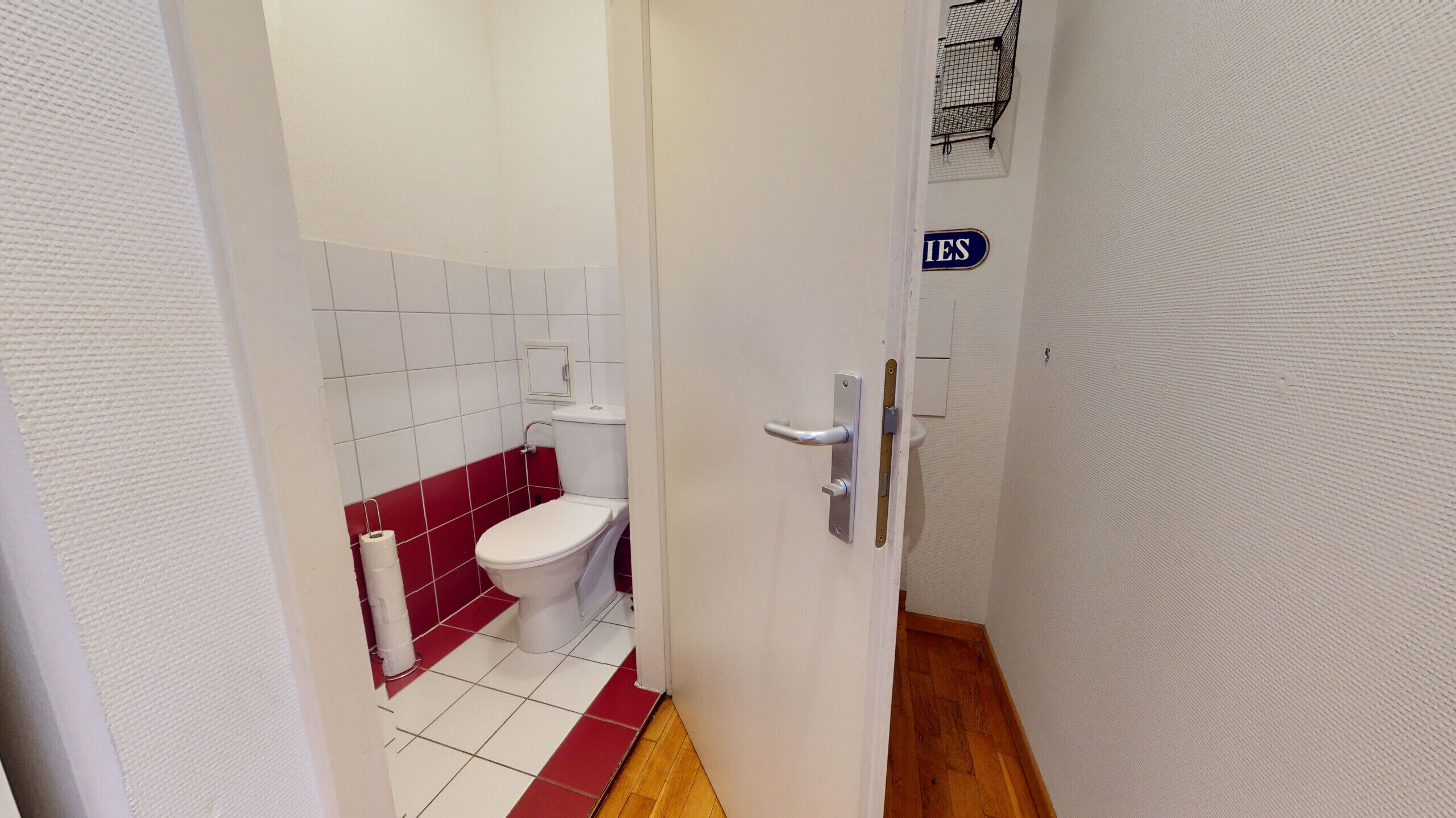 Un couloir étroit mène à une salle de bain équipée de toilettes, dont le sol est recouvert de carreaux rouges et qui possède une porte blanche avec une poignée.