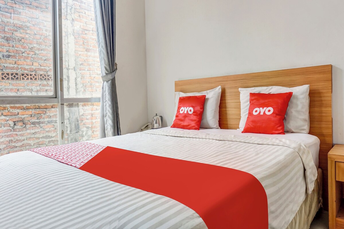 Une chambre d’hôtel avec un grand lit, deux oreillers rouges ornés du texte « OYO », une tête de lit en bois et une fenêtre donnant sur un mur de briques.