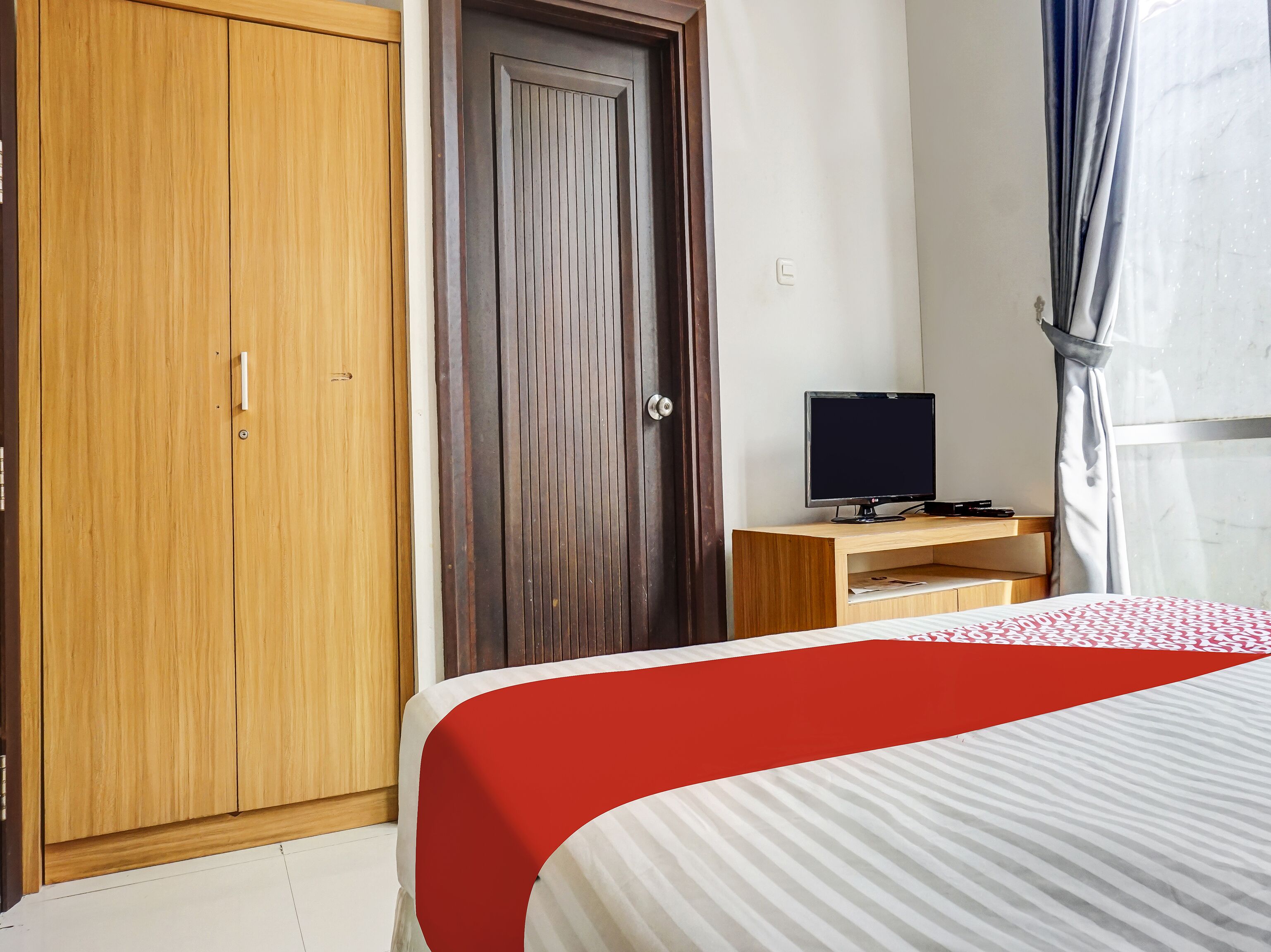 A hotel room with a wooden wardrobe, a closed door, a television on a wooden stand, and a bed with a striped bedspread.