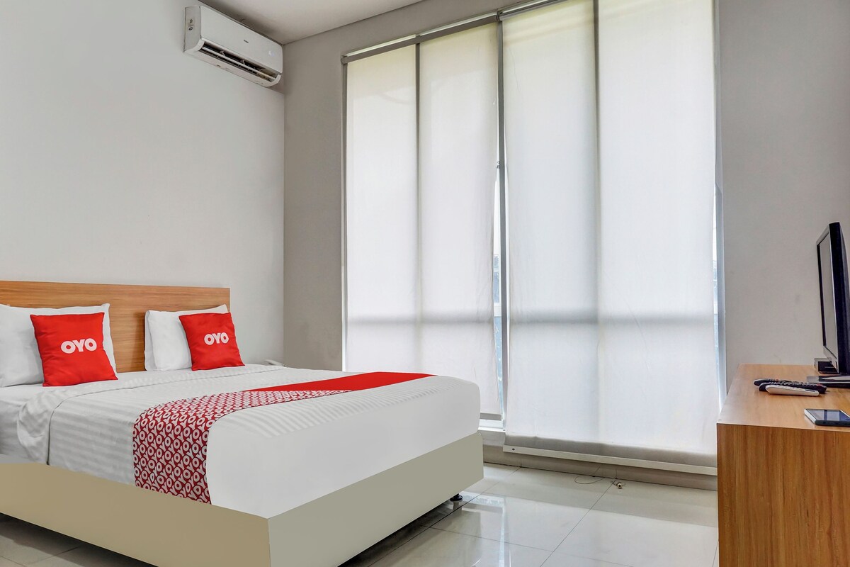 Una cama bien hecha con una colcha estampada, dos almohadas con la palabra “OYO” y una mesita de noche con un control remoto y un teléfono inteligente.