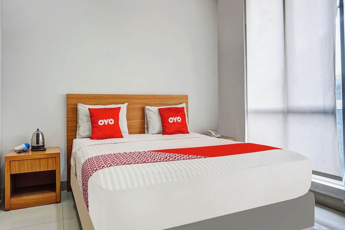 Habitación de hotel con cabecera de madera, dos almohadas rojas con la marca 'OYO', una colcha blanca, una mesita de noche con hervidor y teléfono, y una ventana con cortinas.