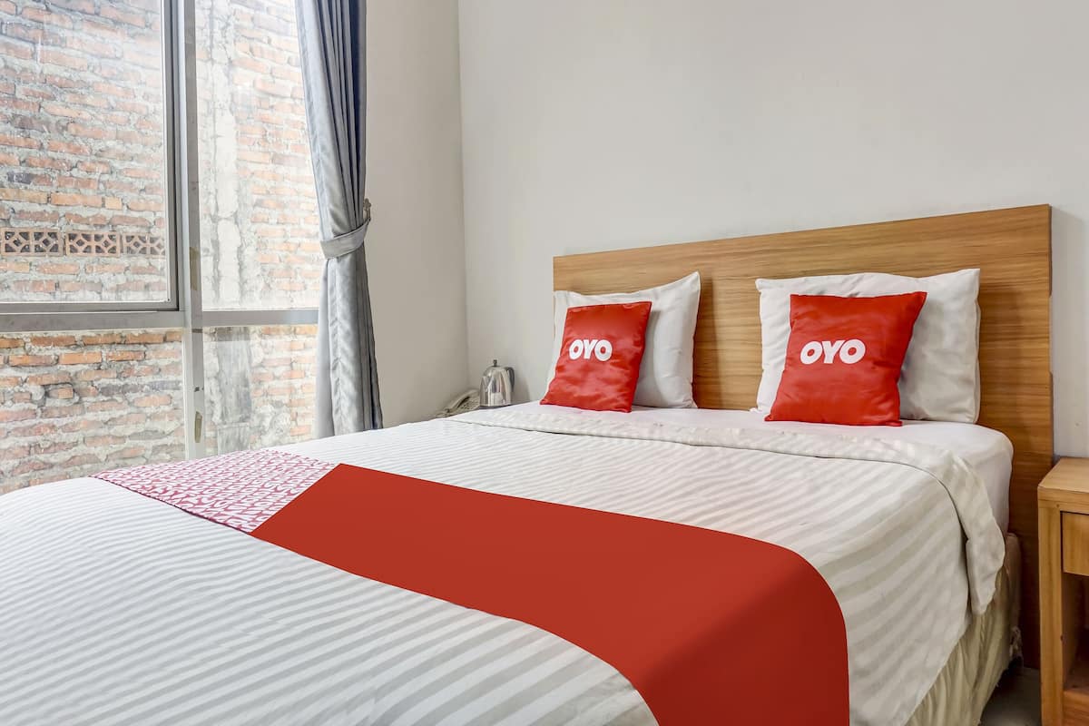 A hotel room with a large bed, two red pillows with "OYO" text, a wooden headboard, and a window with a view of a brick wall.
