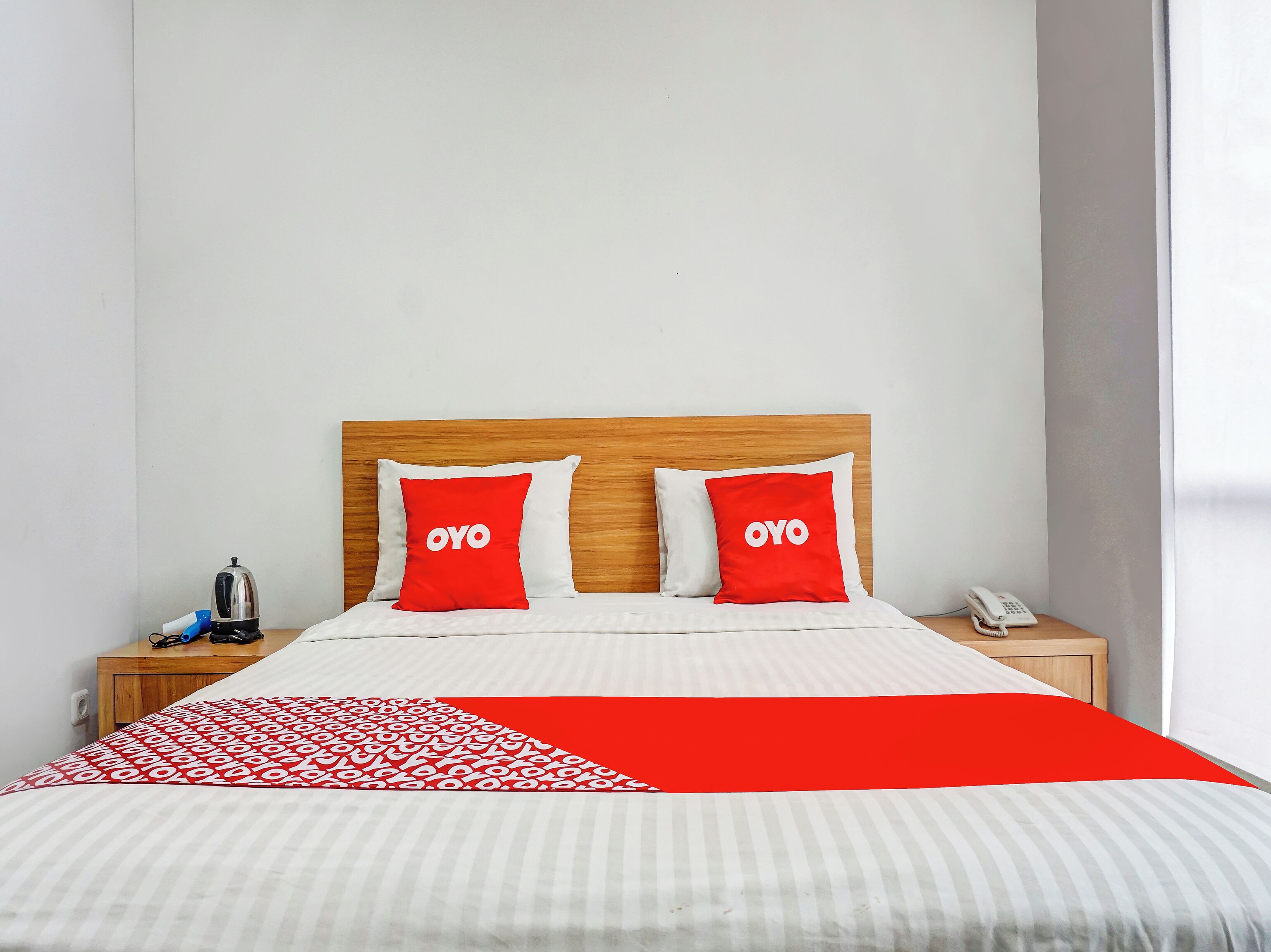 Ein Bett mit zwei roten Kissen, einem Holzkopfteil, einer weiß-rot gestreiften Bettdecke, einem Nachttisch mit Telefon und einem Wasserkocher.