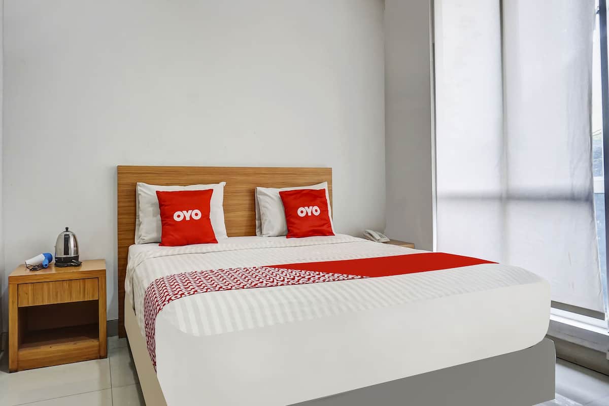 A hotel room with a wooden headboard, two red pillows with "OYO" branding, a white bedspread, a bedside table with a kettle and a phone, and a window with curtains.