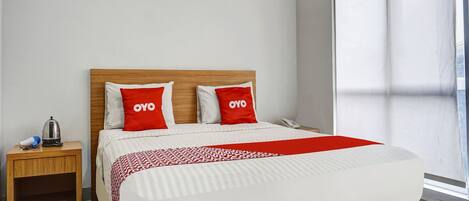 A hotel room with a wooden headboard, two red pillows with "OYO" branding, a white bedspread, a bedside table with a kettle and a phone, and a window with curtains.