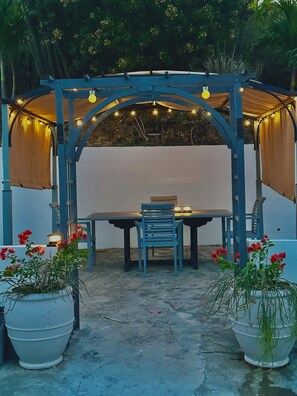 Un espacio al aire libre con techo para comer, que incluye una mesa, sillas, luces colgantes y plantas en macetas.