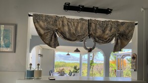 Una habitación con vistas a montañas y vegetación, una ventana con cortinas, un estante con botellas y un teléfono, y una lámpara colgante.