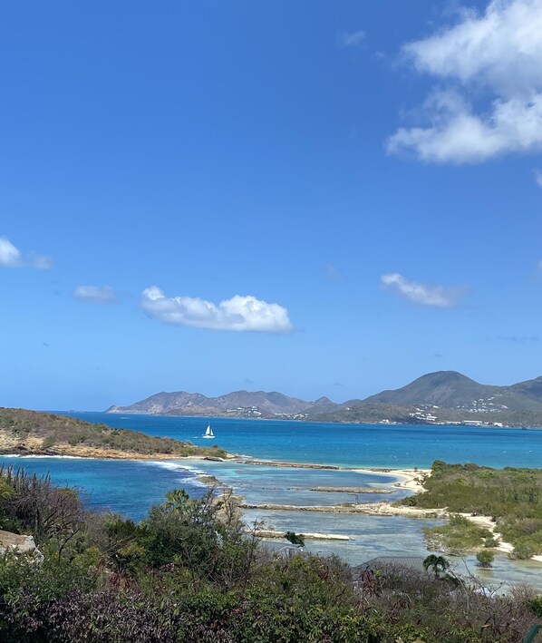 Un paisaje costero con cielo azul despejado, un velero en el agua y montañas a lo lejos.