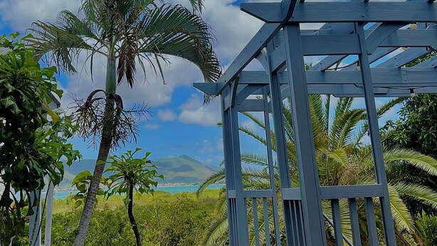 Una estructura de pérgola de madera con vistas a un paisaje costero, que incluye palmeras y una montaña distante.