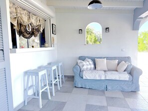 Une pièce avec un canapé bleu clair, des tabourets de bar blancs, une fenêtre donnant sur un paysage verdoyant et un ventilateur de plafond.