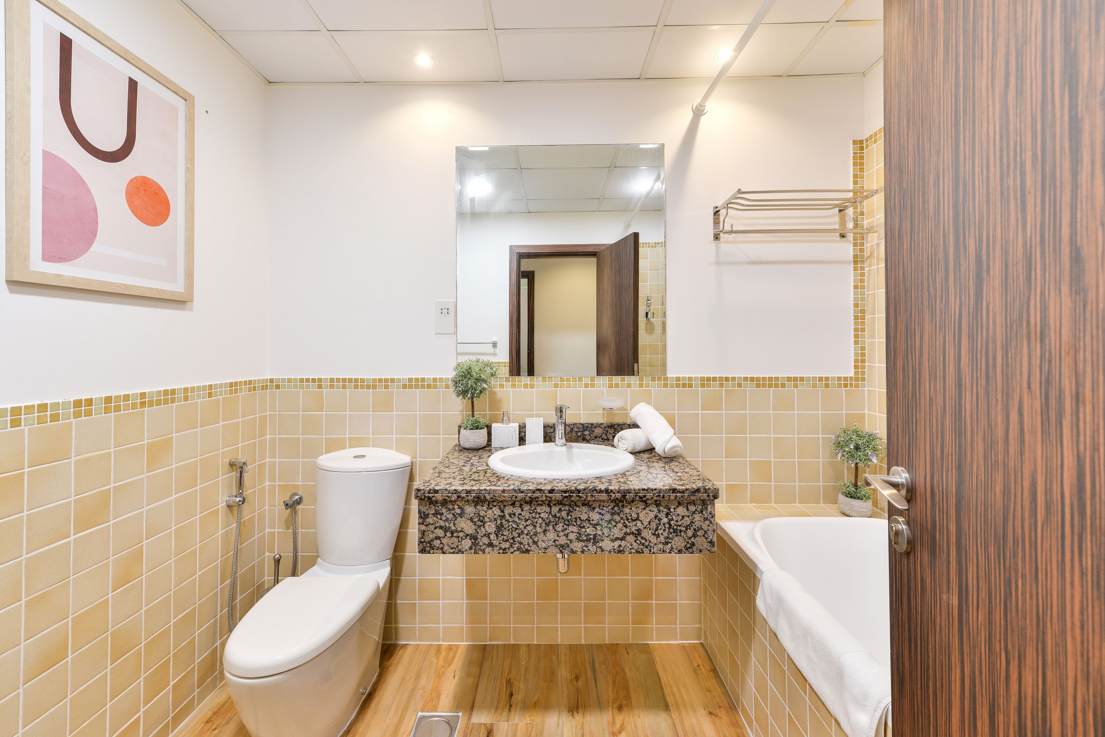 A bathroom with a toilet, sink, and bathtub, tiled in beige and brown, with a wooden door and a mirror above the sink.