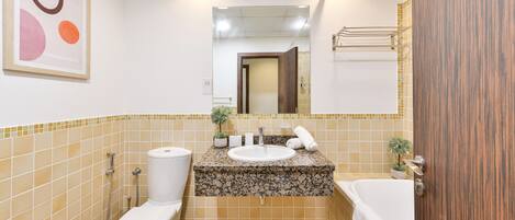 A bathroom with a toilet, sink, and bathtub, tiled in beige and brown, with a wooden door and a mirror above the sink.
