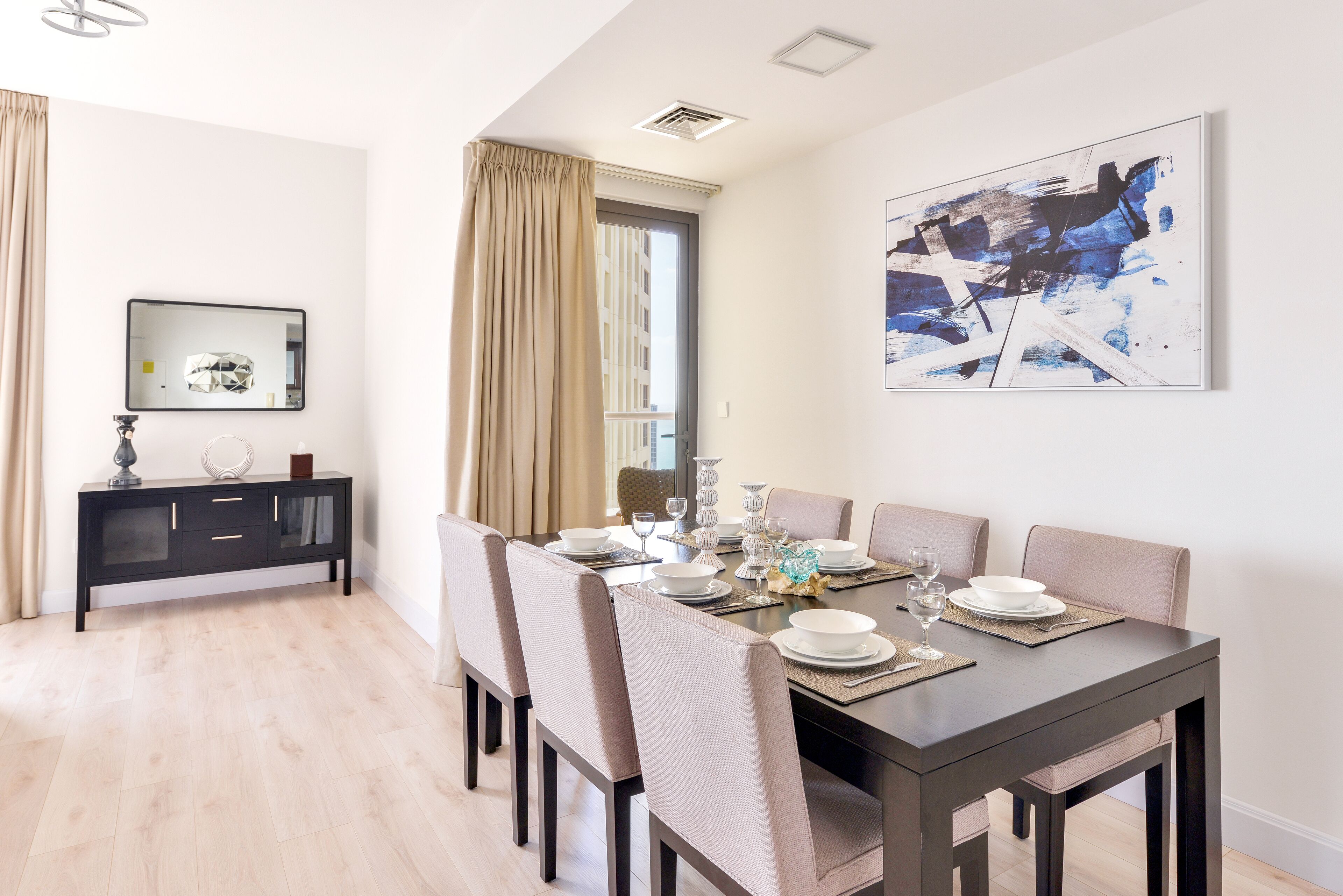 Een eetruimte met een donker houten tafel voor zes personen, omringd door beige stoelen. Een zwart console tafeltje met een spiegel en decoratieve objecten staat tegen de muur. Daarboven hangt een groot abstract schilderij.