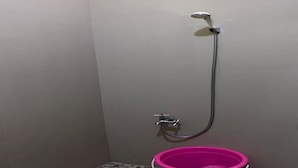 Un espace douche avec un mur recouvert de carreaux gris, une pomme de douche et un seau rose.
